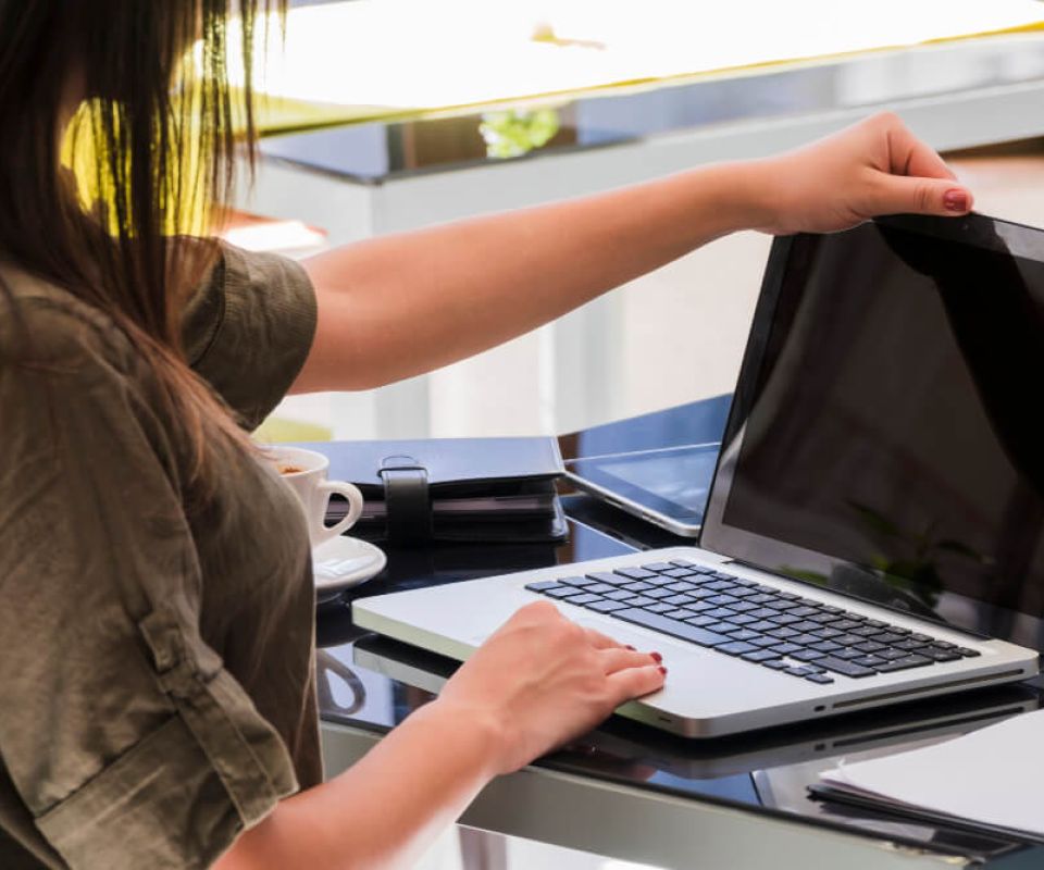 Person opening a laptop on a desk in a modern office workspace, with a coffee cup, notebooks, and a small plant nearby.