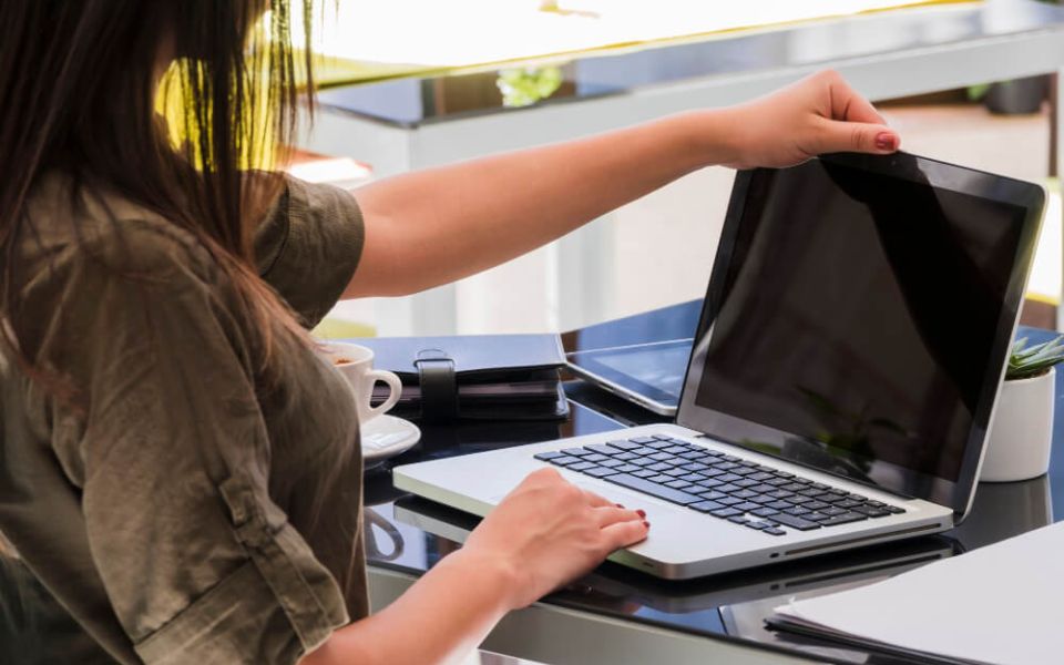 Person opening a laptop on a desk in a modern office workspace, with a coffee cup, notebooks, and a small plant nearby.