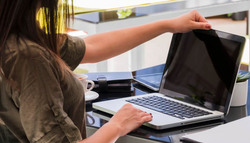 Person opening a laptop on a desk in a modern office workspace, with a coffee cup, notebooks, and a small plant nearby.
