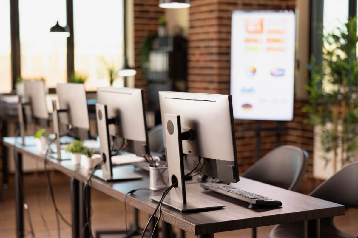 Modern open-plan office workspace with a row of desktop computer monitors on long wooden desks, keyboards and office supplies neatly arranged