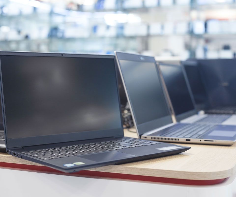 A row of modern laptops displayed on a wooden table in an electronics store, with several open devices and a blurred retail showroom in the background.