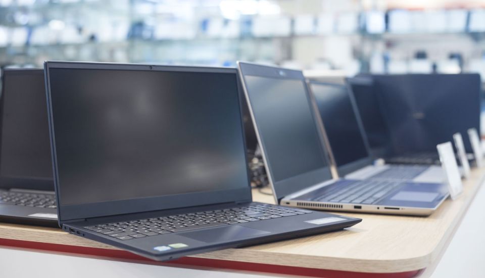 A row of modern laptops displayed on a wooden table in an electronics store, with several open devices and a blurred retail showroom in the background.