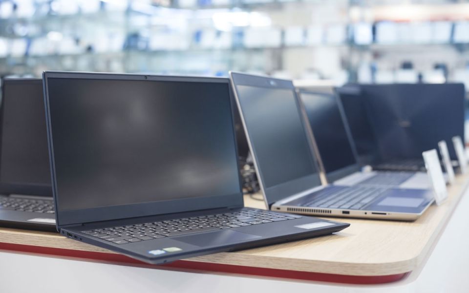 A row of modern laptops displayed on a wooden table in an electronics store, with several open devices and a blurred retail showroom in the background.