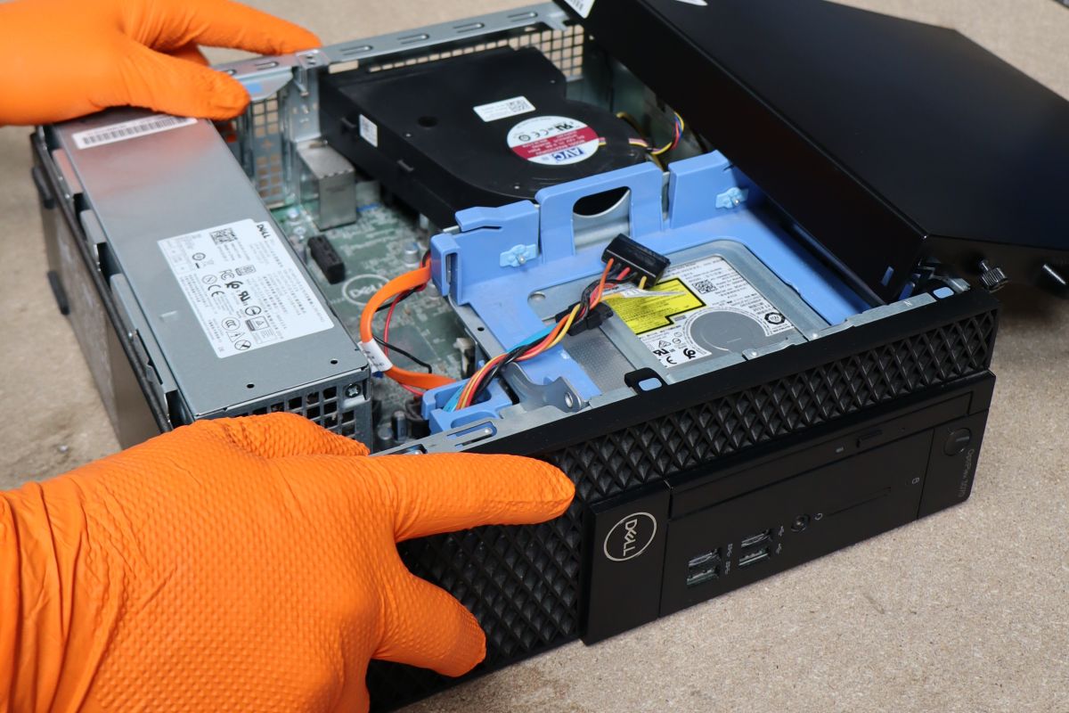 A person wearing bright orange protective gloves opens a small Dell desktop computer case, revealing internal components including the power supply, cooling fan, hard drive, and cables during maintenance or repair.